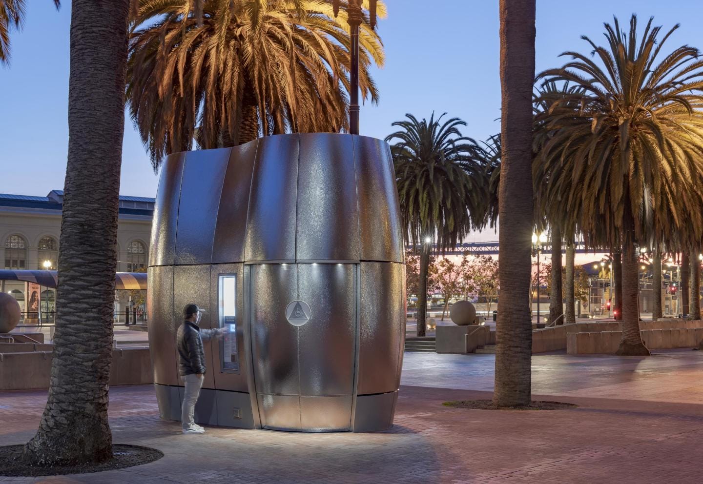 A person standing in front of a futuristic-looking silver public toilet kiosk nestled among palm trees in San Francisco's Embarcadero plaza