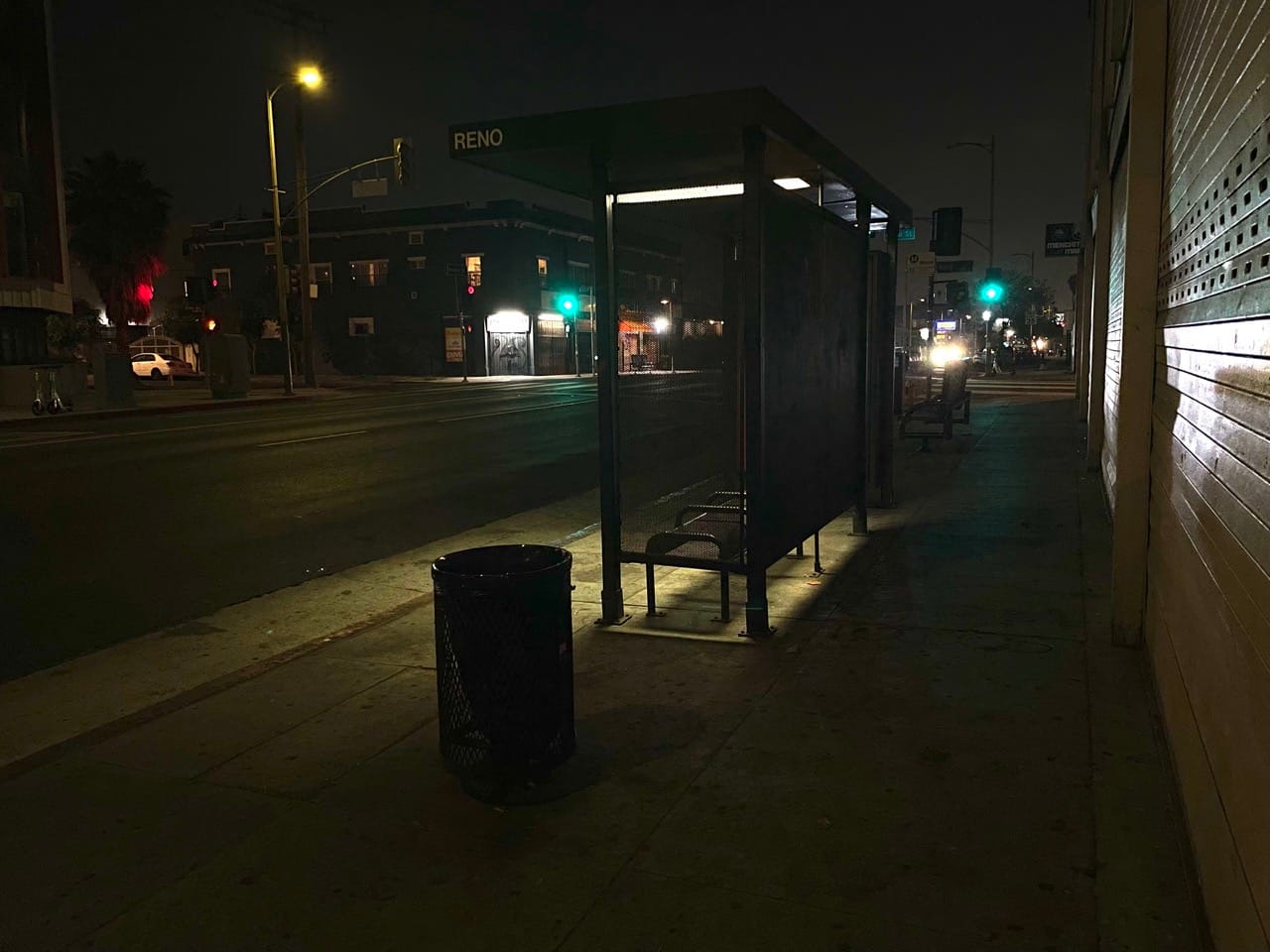 A very dark bus stop on an empty street with a bus shelter providing the only light. Two streetlights are dark as well