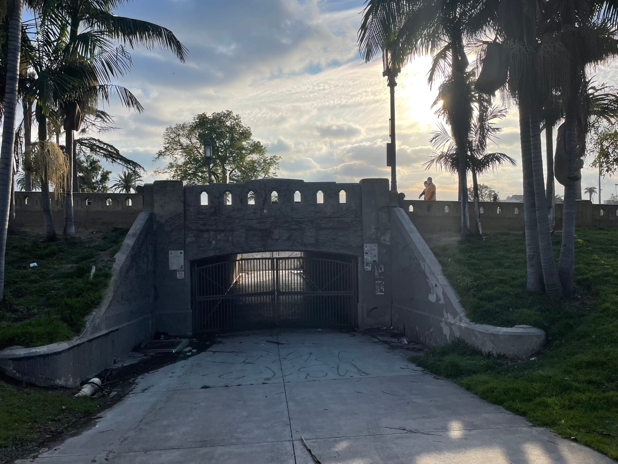 A pedestrian bridge under a roadway in MacArthur Park, with grassy hillsides and palm trees and two people walking on the road above