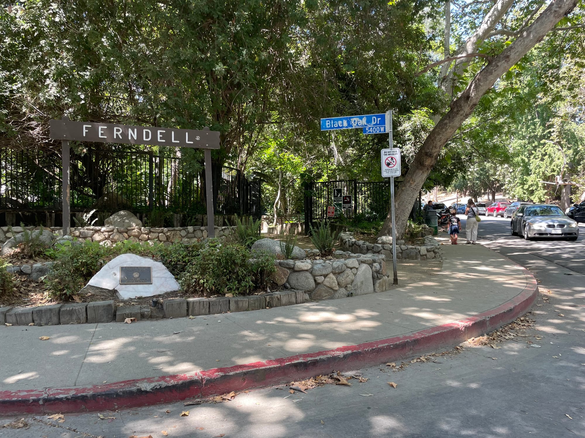 A shady forest in Griffith Park's Ferndell nature path, with a Ferndell sign and a river rock gate, fronted by a bright red curb and no ramp to access it from the street