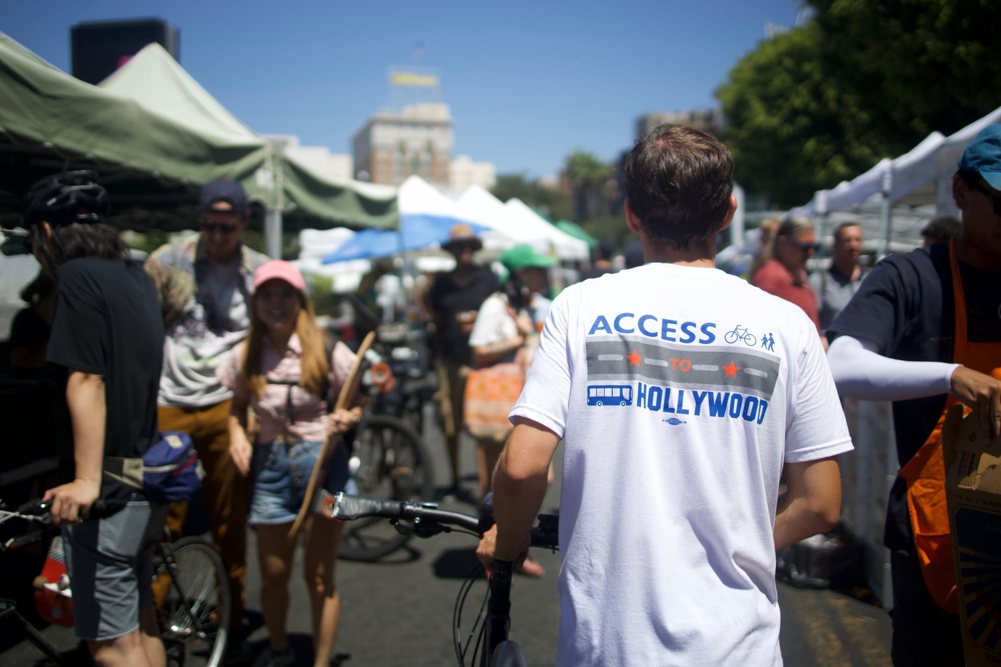 A man wearing an "access to hollywood" shirt showing bikes, pedestrians, and buses on Hollywood boulevard as he walks around a busy farmers market