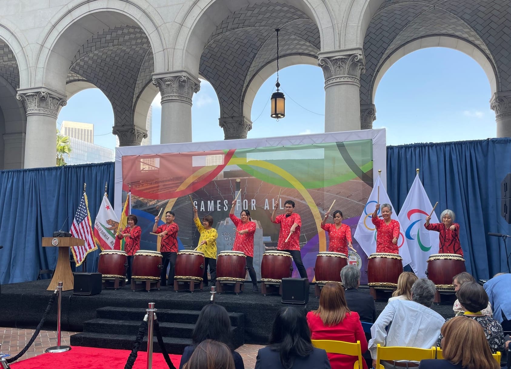 Taiko drummers dressed bright red and yellow tops bang huge drums on a stage that says Games for All at LA's city hall