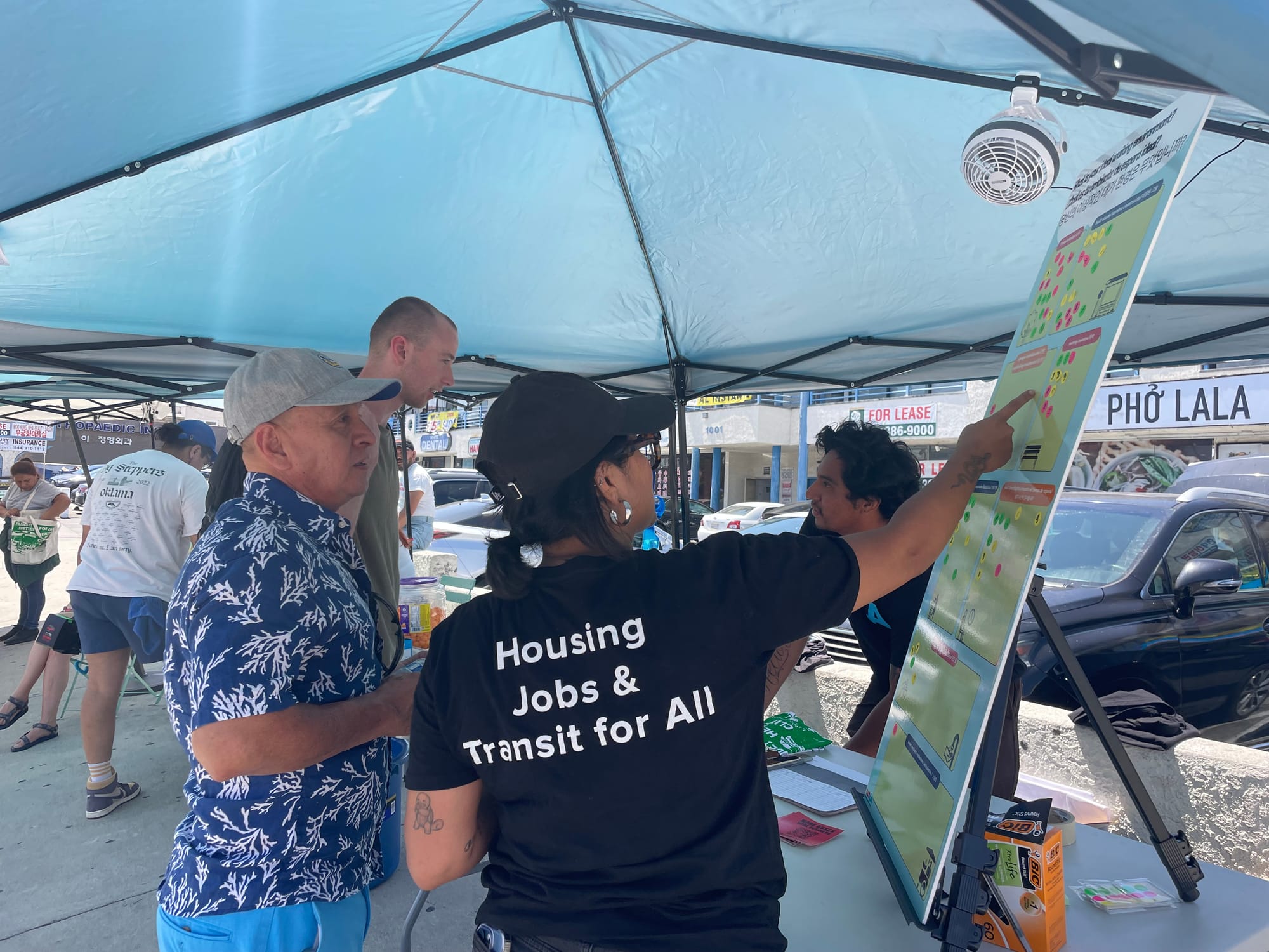 A person with a shirt that says housing jobs and transit for all helps a passenger put stickers on a board