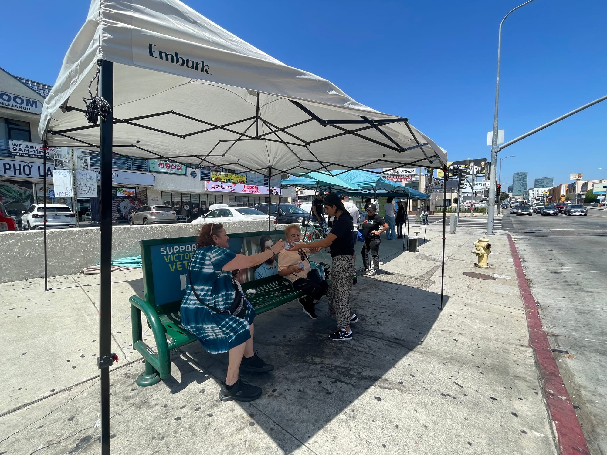 People pass out waters and snacks under a pop up tent at a bus bench on a busy street