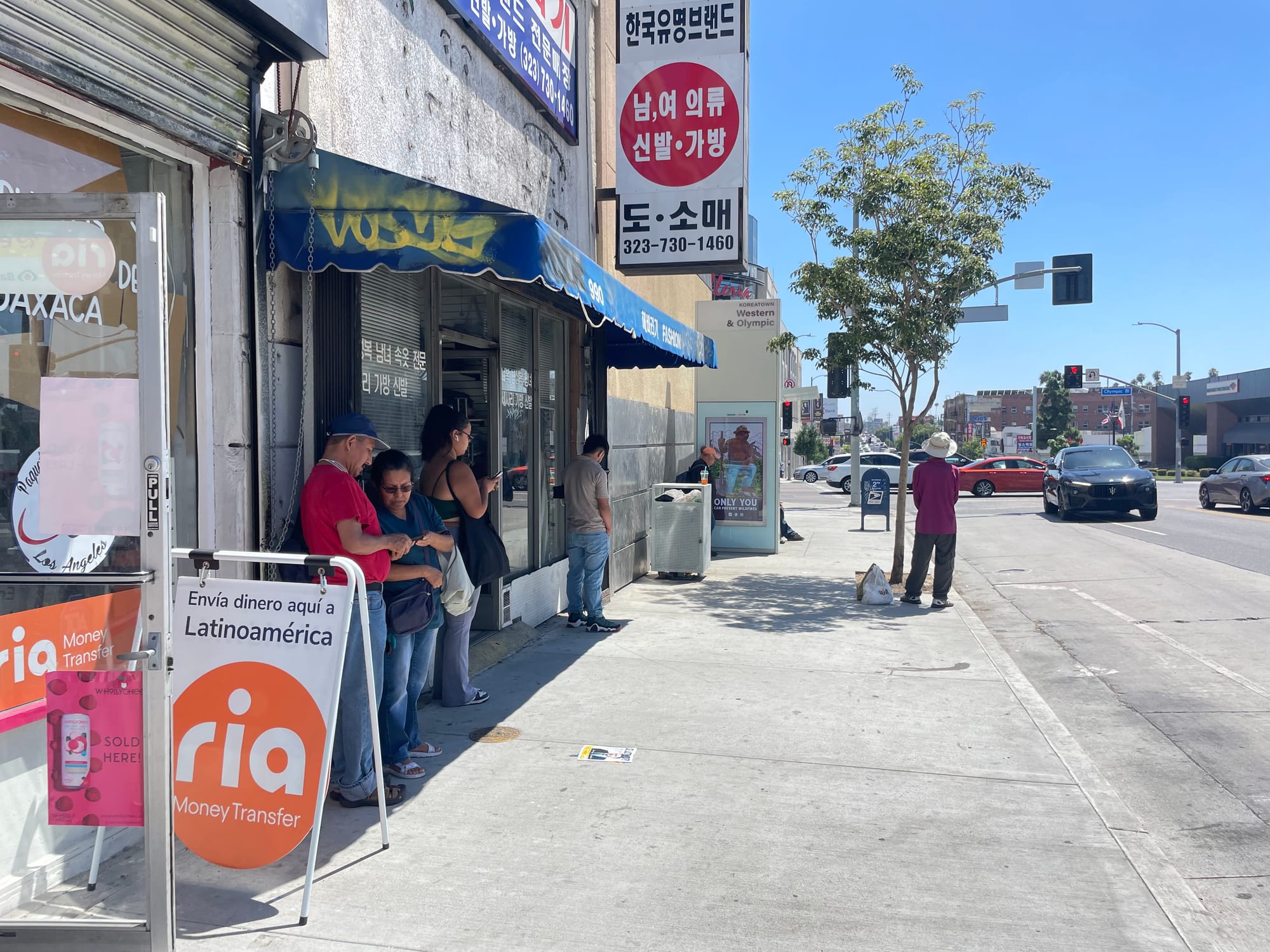 People waiting for the bus at a shadeless stop huddled into the shade along the buildings