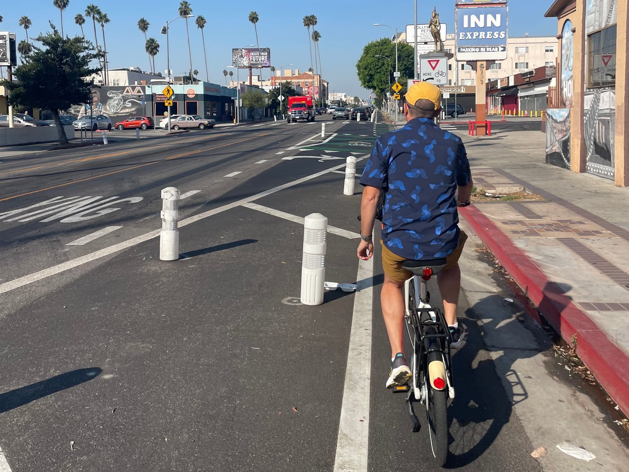 A man in a blue shirt and yellow hat riding a bike share bike down a bike lane on Hollywood Boulevard lined with white bollards