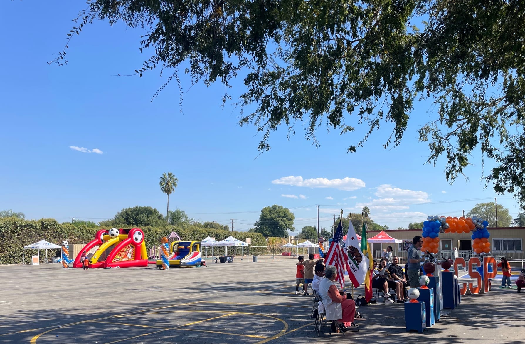 A press conference for the community school park program with balloons and speakers at a podium taking place in a large asphalt schoolyard