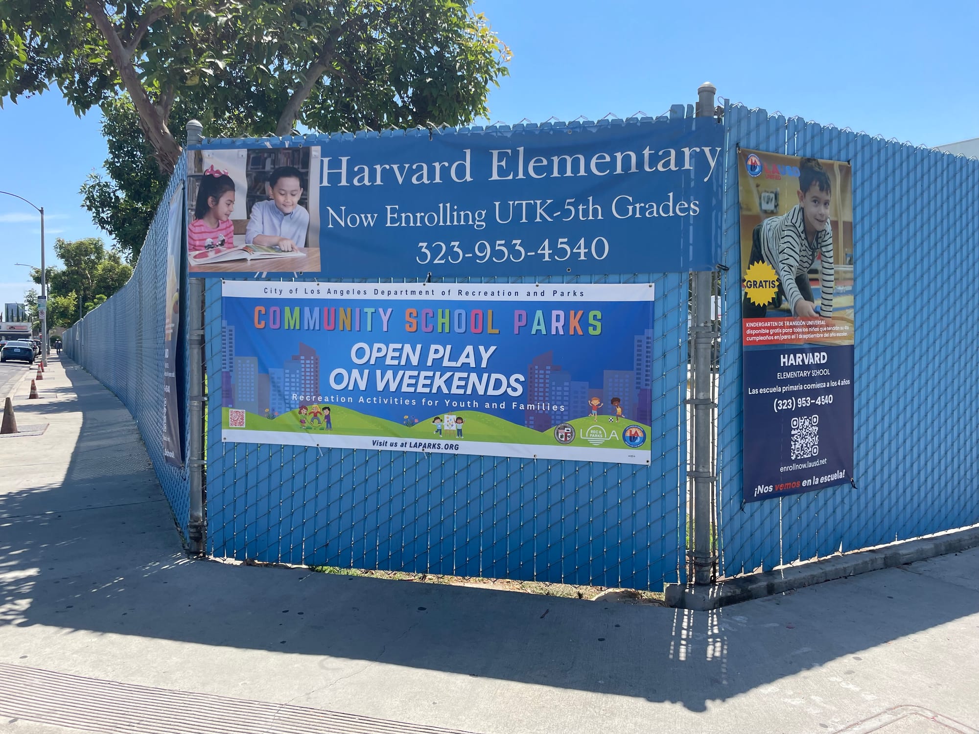 A blue fence with signs that identify it as Harvard elementary school, with a large banner that says Community School Parks, Open Play on Weekends