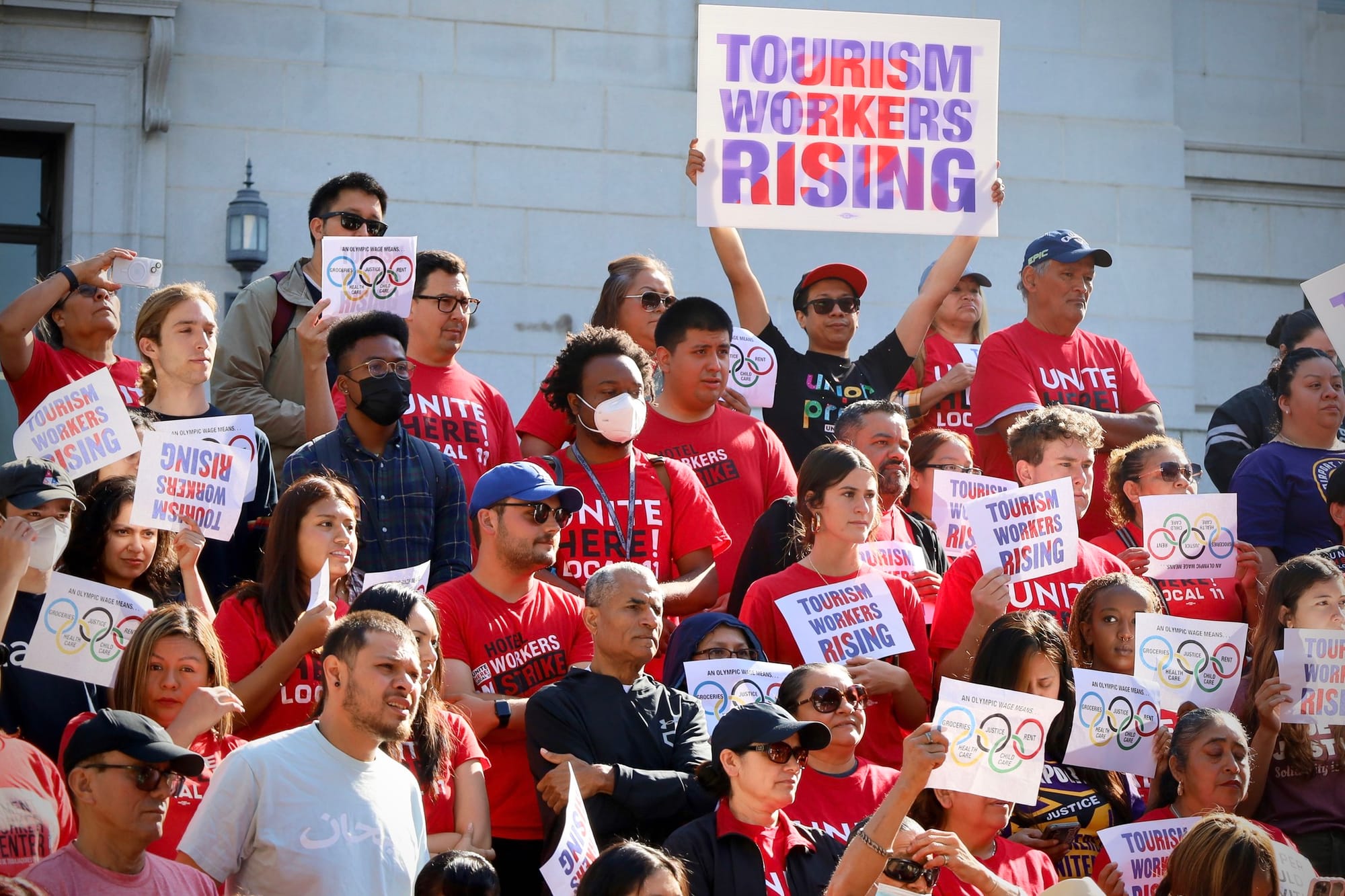 A crowd on the steps of City Hall wearing Unite Here red shirts and holding signs that read: tourism workers rising