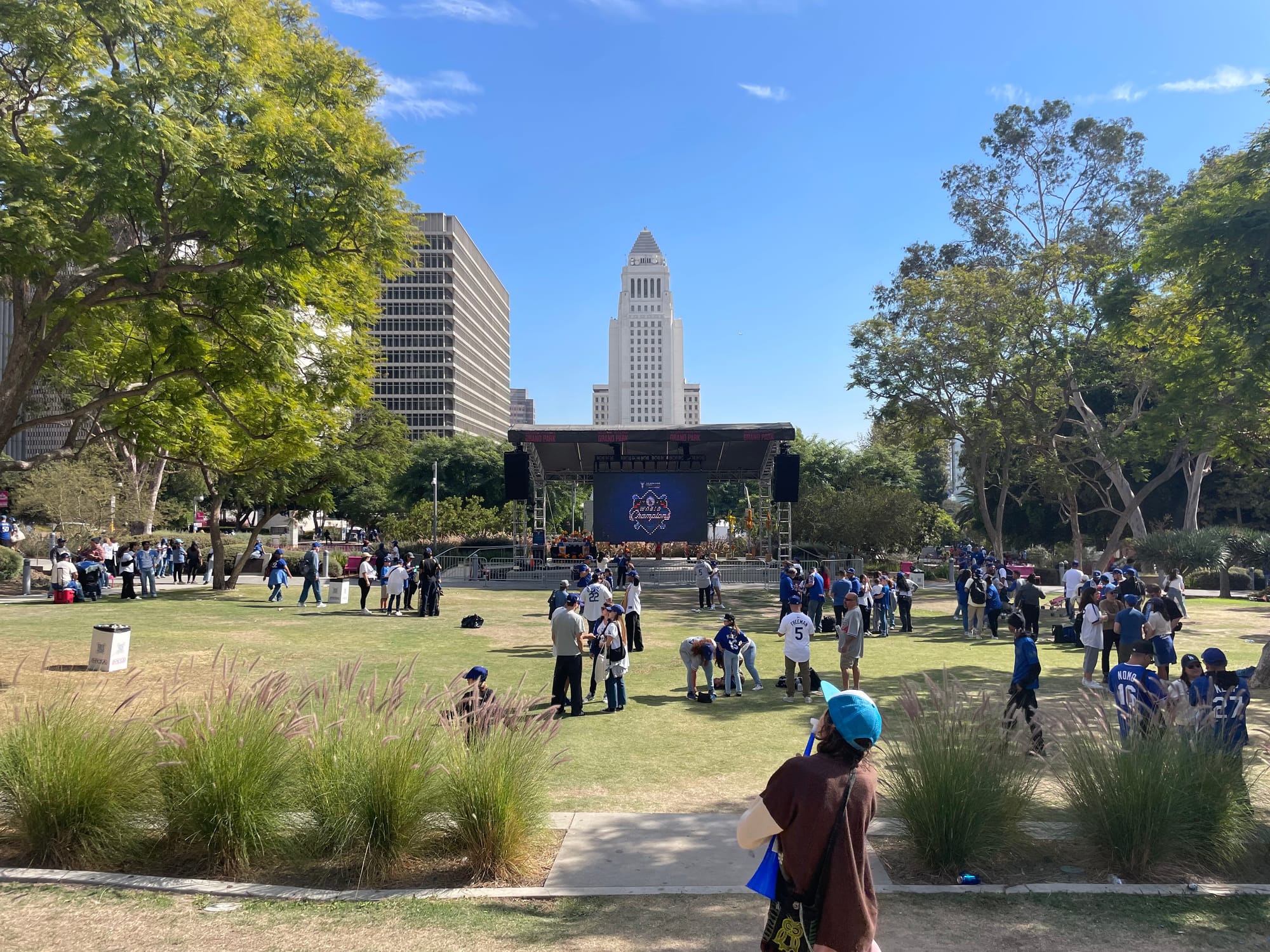 Dodger fans wearing blue and white mill about Grand Park in a grassy area with City Hall in the background