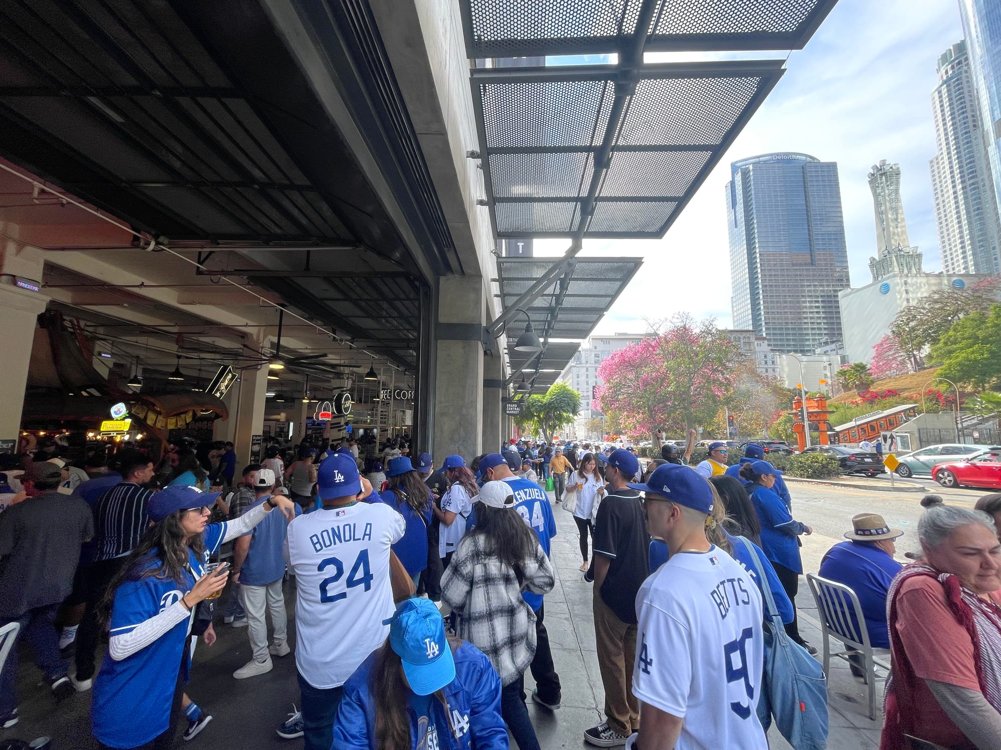 Dodger fans filling the sidewalk in front of Grand Central Market's food hall with the towers of downtown in the background