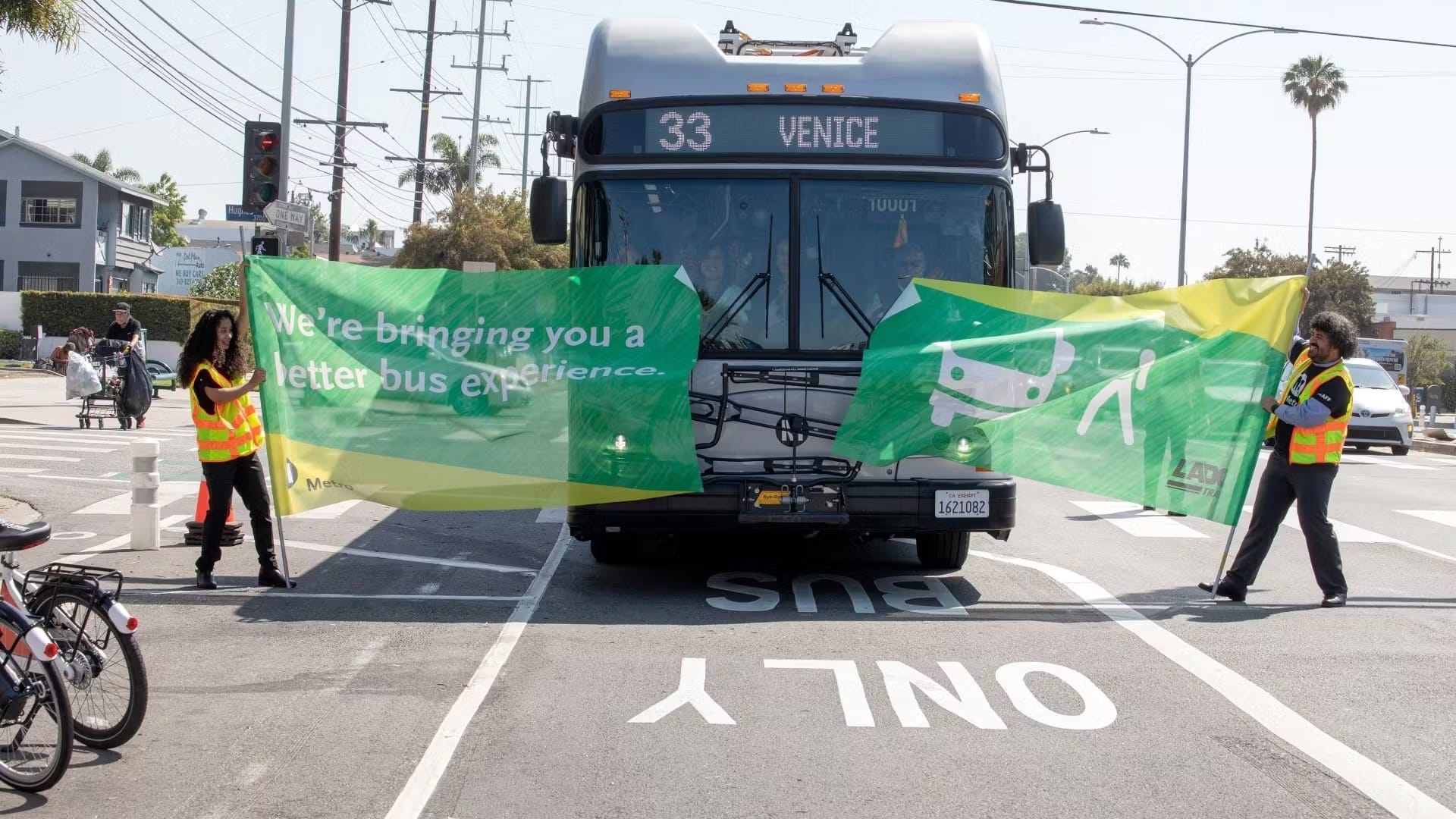 A silver Metro bus with the headsign reading VENICE is breaking through a green banner held by two workers in safety vests that says we're bringing you a better bus experience! the bus is traveling in a bus only lane