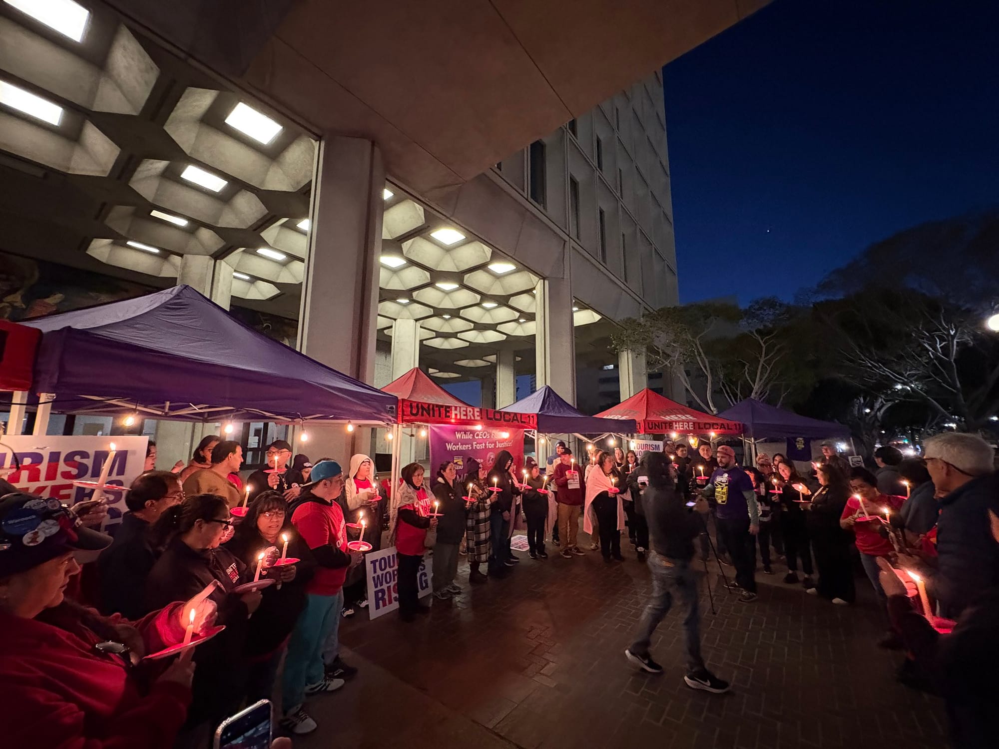 Tourism workers in a candlelight vigil outside City Hall wearing red and purple shirts and holding candles and signs that say Unite Here and Tourism Workers Rising