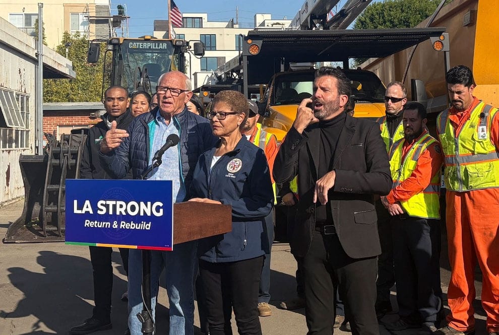 Behind a podium that says LA STRONG return and rebuild, Mayor Karen Bass stands with Steve Soboroff and construction equipment