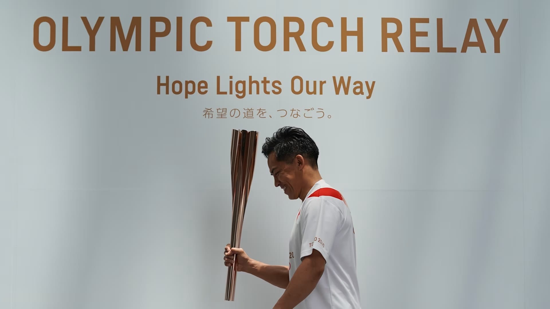 An athlete walking the gold torch along a wall that reads Olympic Torch Relay, hope lights our way