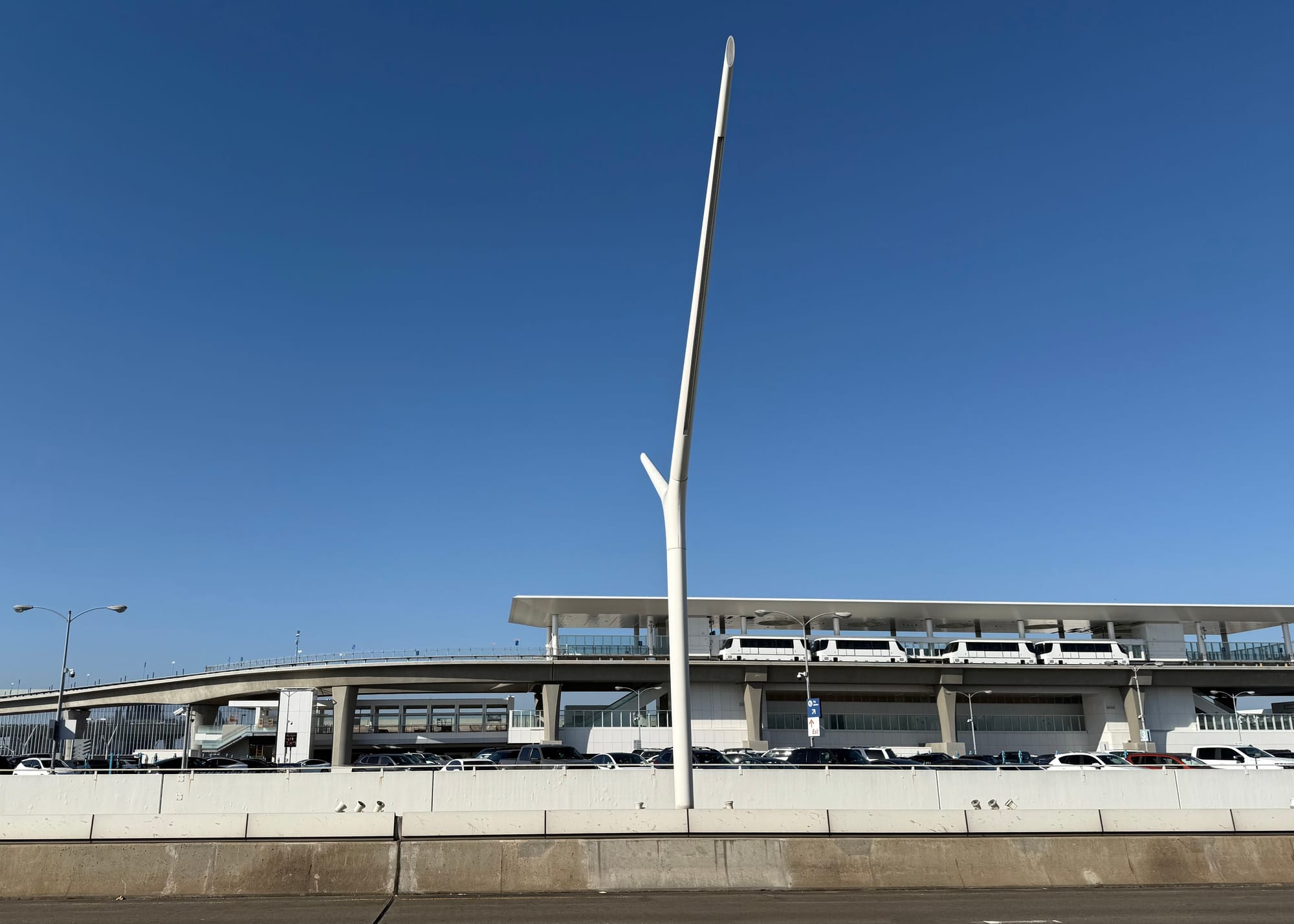 The LAX people mover is in action with four large white tram cars parked at an elevated station with the parking lots of the airport all around it